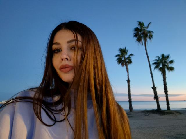 ValeriMair with long brown hair, full lips, and a serious expression, wearing a white shirt, standing in front of palm trees at sunset. Her tits are small and her pussy is not visible.