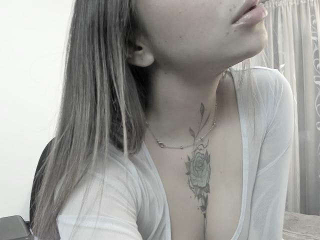 KayleeM's face tilted up, showing her tits and a rose tattoo on her chest. She's wearing a sheer white top, with long grayish-brown hair. Necklace around her neck.