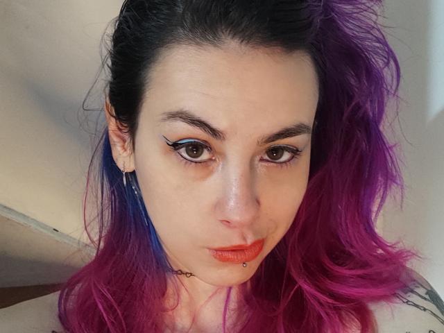 NatashaLau has purple and blue hair, brown eyes with black eyeliner, and a lip piercing. Her face is centered, showing a serious expression. She's wearing a choker.