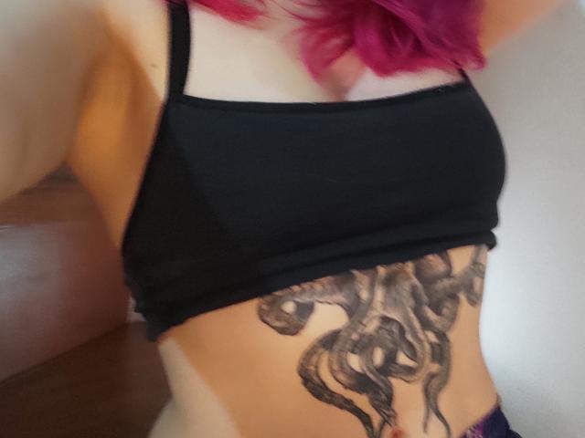 NatashaLau with pink hair, black tank top, and a large octopus tattoo on her lower stomach, showing her tan lines. Her tits are small, her pussy is not visible.