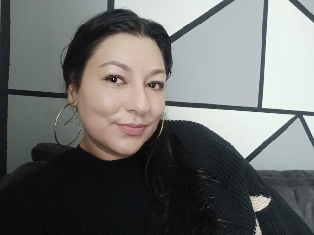 SakotoDika has medium skin, black hair, and is wearing a black, ripped, knit sweater with large hoop earrings. She's smiling with pink lipstick, brown eyes.