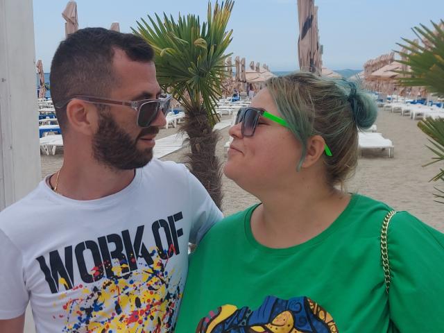 Nikole ha l'ombretto verde, una maglietta verde e i capelli raccolti in uno chignon, ed è in piedi accanto a Jim, che ha la barba, indossa una camicia bianca e occhiali da sole, su una spiaggia..