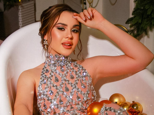 EricaWings draagt een zilveren top, versierd met edelstenen, en zit in een wit bad met gouden en oranje versieringen. Ze legt haar hand op haar hoofd en kijkt met een lichte glimlach naar de camera..