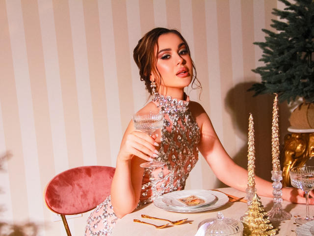 EricaWings mit einem glitzernden Kleid, die ein Glas hält, sitzt auf einem rosa Samtstuhl, vor ihr ein Tisch mit goldenen Kerzen und ein Weihnachtsbaum. Sie schaut mit einem leichten Lächeln in die Kamera, trägt glänzende Ohrringe und hat eine hochgesteckte Frisur..