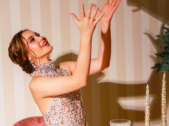 EricaWings, mit erhobenen Armen, lächelnd, trägt ein glitzerndes, hochgeschlossenes, ärmelloses Kleid, wobei ihre Brüste leicht sichtbar sind, braune Haare zu einem Dutt gebunden, rote Lippenstift, steht in der Nähe eines rosa Stuhls..