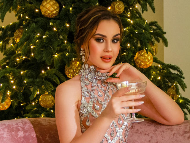EricaWings, vestindo um vestido prateado brilhante, segurando uma taça de champanhe, usando brincos pendentes, em frente a uma árvore de Natal decorada, parecendo adorável com olhos castanhos, lábios rosados e cabelo castanho ondulado..