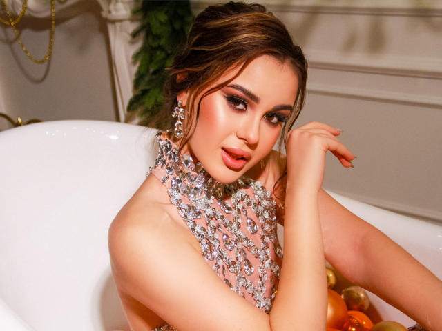 EricaWings with shiny tits in a white bathtub, wearing a sparkling, jeweled necklace, looking at the camera with a sexy, open-mouthed smile, brown hair in a messy bun.
