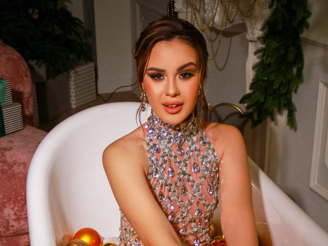 EricaWings trägt ein sexy, durchsichtiges, silbernes Kleid mit Pailletten und glitzernden Schmucksteinen und sitzt in einer weißen Badewanne mit orangefarbenen Dekorationen. Sie wirkt verführerisch mit ihren dunklen, welligen Haaren und dem strahlenden Make-up..