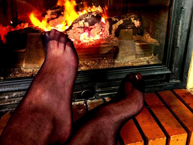 Les pieds marron foncé de LilouLove, avec des soles légèrement poilues, les orteils pointant vers le bas, reposent sur un sol en briques devant une cheminée où brûlent des flammes orange et jaunes vives..