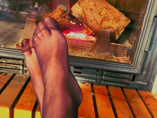 LilouLove's feet, with slightly dirty toes, resting on a wooden floor in front of a burning fireplace, showing a piece of burning wood inside the glass.