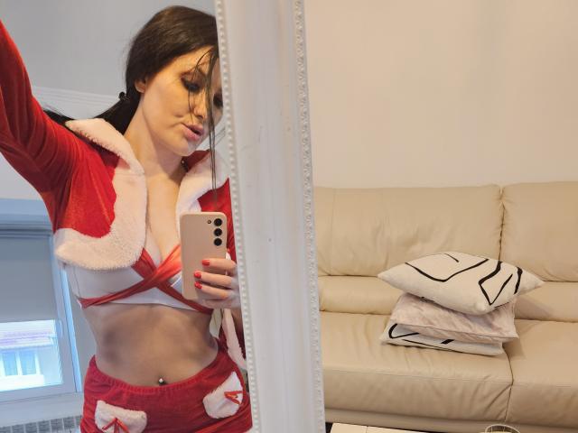 JoliDanielle, vestita con un costume rosso da Babbo Natale con bordi di pelliccia bianca, mostra i suoi seni in un selfie allo specchio, tenendo un telefono, con le unghie rosse, e un divano bianco sullo sfondo..
