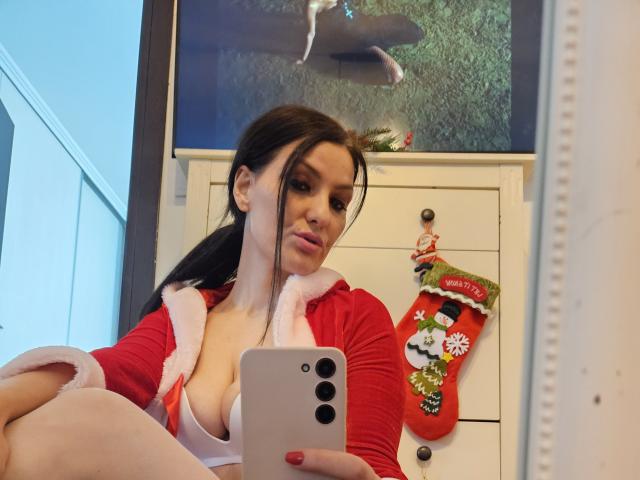 JoliDanielle with dark hair, red Santa jacket, white fur trim, and exposed large tits, holding a silver iPhone, taking a selfie in a mirror, Christmas stocking in background.