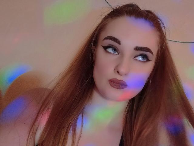 KariMillie, met lang rood haar, dikke wenkbrauwen, blauwe ogen en paarse lippenstift, kijkt opzij en heeft kleurrijke, regenboogkleurige lichtreflecties op haar gezicht en borst..