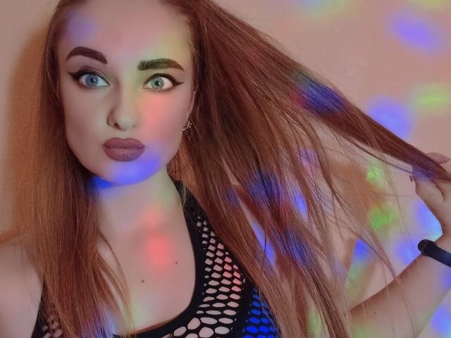 KariMillie, com olhos azuis, sobrancelhas escuras e cabelo ruivo, segurando o cabelo, vestindo uma blusa de malha preta, reflexos de luz coloridos no rosto e uma pulseira preta..