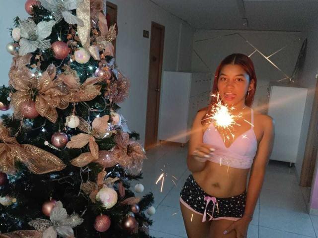 HilaryCook, avec des cheveux roux, un soutien-gorge rose et un short noir à pois, tenant une allumette, debout à côté d'un sapin de Noël décoré, souriant les yeux fermés..