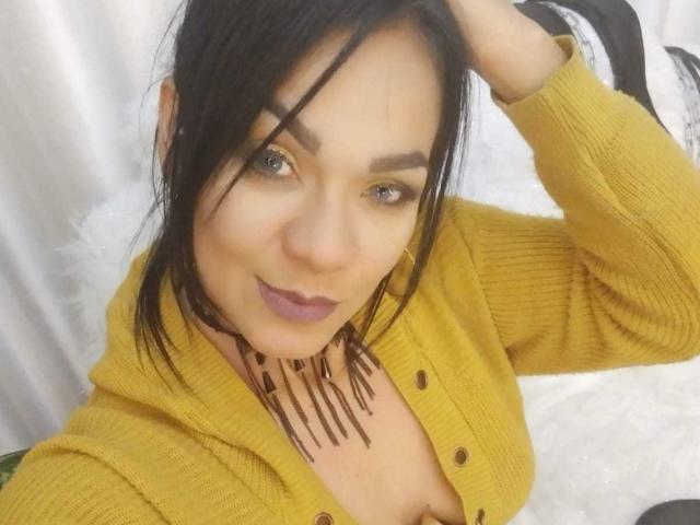 CharlotteJhons, avec ses cheveux noirs, son cardigan jaune et son collier marron, dévoilant sa poitrine, avec du rouge à lèvres violet et un léger sourire, une main dans les cheveux, regardant l'appareil photo..