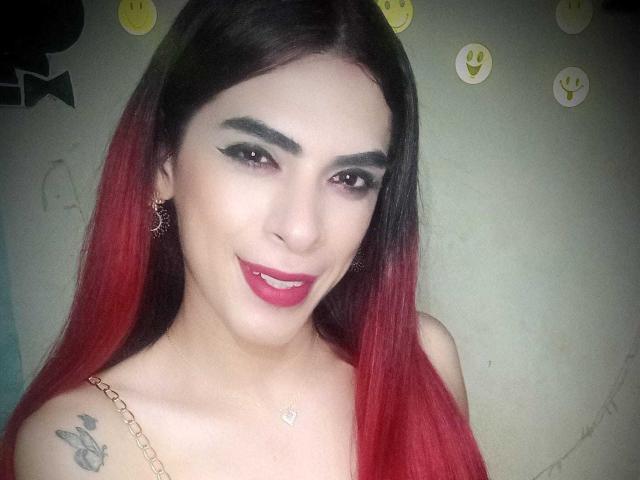 KataleyaHoty, con capelli rossi e neri, sopracciglia nere folte, rossetto rosso, orecchini e un tatuaggio sulla spalla destra, sorride alla fotocamera. Indossa una collana..