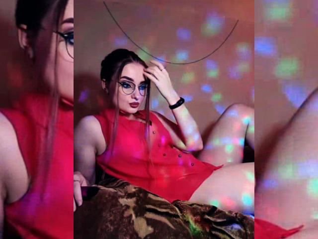 KariMillie, avec des lunettes, une robe rouge et des cheveux foncés, assise les jambes écartées, avec des reflets de lumière colorés, une main dans les cheveux, un bracelet noir, une couverture marron, et des sous-vêtements rouges visibles..