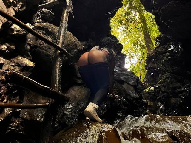 Amberv's ass is exposed, bent over with blue jeans pulled down, black leggings, and white socks in a rocky, dark forest with a bright green leaf above.