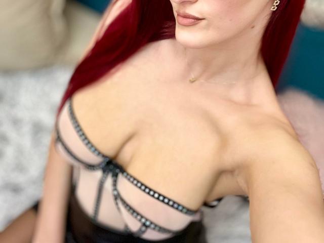 Rosalyn with red hair, wearing a sheer black bra with straps, showcasing her tits, pale skin, and a small gold necklace, taken from a slightly above angle.