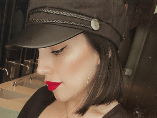 Liliysweet with dark bob, black cap, red lipstick, thick black eyeliner, large hoop earrings, pale skin, and a slight smile. Boobs partially visible, black top, subtle makeup.