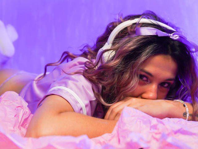 HellenK with wavy brown hair, lying on pink bed, wearing white headphones and pink shirt, face in hands, purple lighting, looking up with sultry eyes.