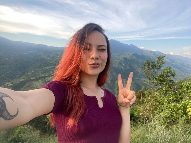 ValeryHarllow with red hair, wearing a purple top, making a peace sign, showing a tattoo on her arm, and smiling at the camera. Sunlight highlights her skin.