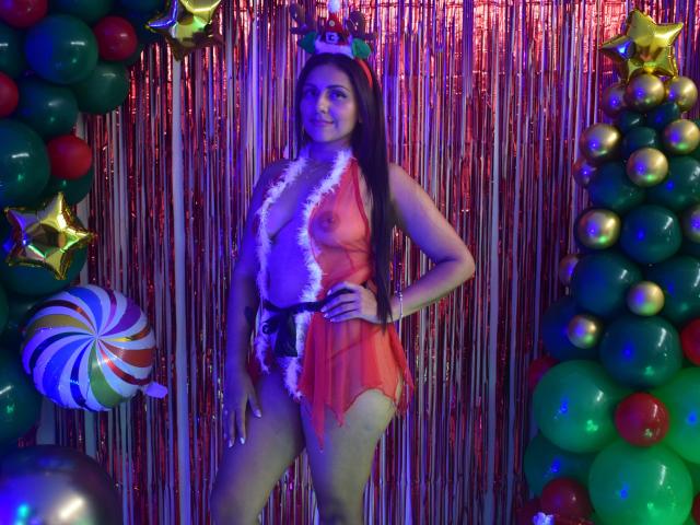 KatyaMore with long brown hair, wearing a red sheer lingerie with white fur trim, exposing her small tits and bare pussy, standing in front of festive balloons and ornaments.