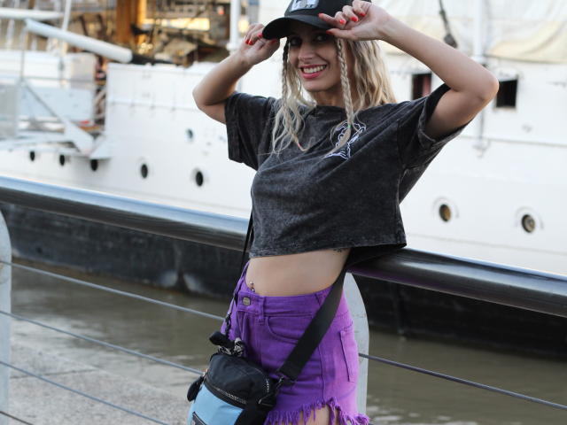 DelaMoons in a dark t-shirt, purple fringed shorts, and a black cap, smiling with hands on cap, showing red nails, and a black cross necklace, standing by a dock.