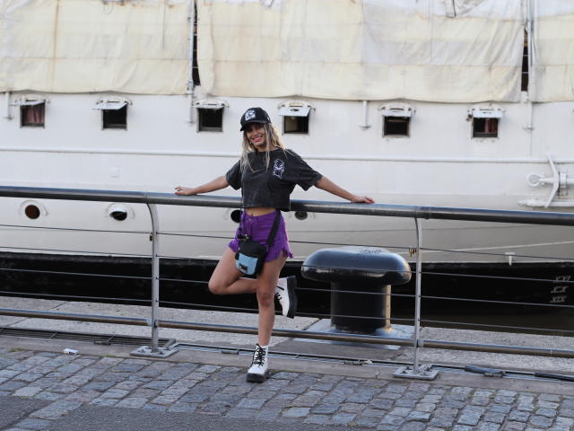 DelaMoons con capelli biondi, cappello nero, top corto grigio, pantaloncini viola, scarpe da ginnastica bianche, appoggiata a una ringhiera di metallo, barca sullo sfondo, pavimento in pietra, una gamba sollevata, sorridente..