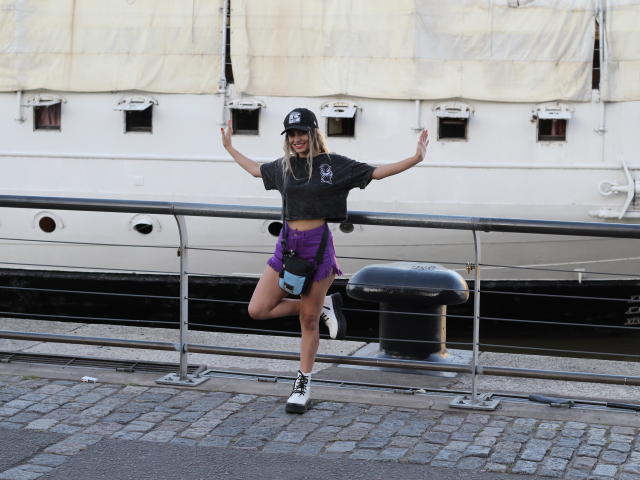 DelaMoons se tient les bras levés, portant une casquette noire, un t-shirt gris, un short violet et des baskets noires, devant un mur blanc d'un navire, avec une jambe pliée au niveau du genou..