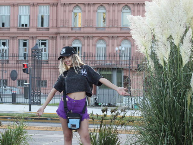 DelaMoons with blonde hair, black cap, grey crop top, purple shorts, and camera strap, standing in front of tall white pampas grass, smiling with arms out.