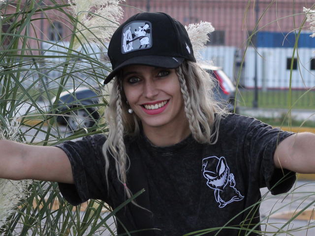 DelaMoons, avec ses tresses blondes, son bonnet noir et sa chemise sombre, sourit les bras ouverts, entourée d'herbes hautes. Elle a du rouge à lèvres rose, un sourire mignon et un t-shirt de groupe de rock..