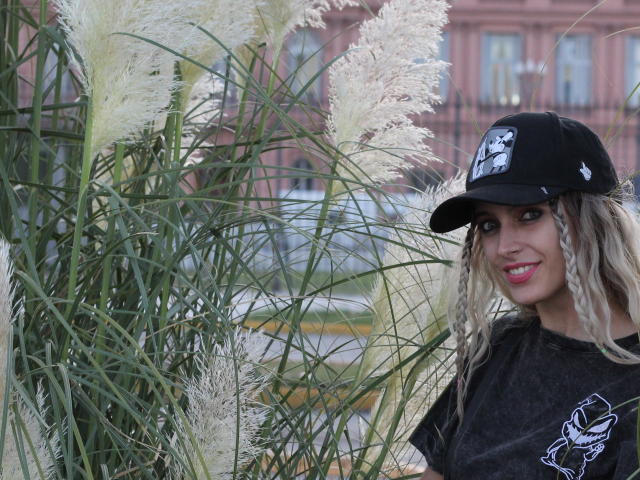 DelaMoons, con largas trenzas rubias, un gorro negro y una camiseta negra, está de pie junto a hierba pampas blanca y esponjosa, sonriendo. Sus senos no son visibles. Es linda y atractiva..