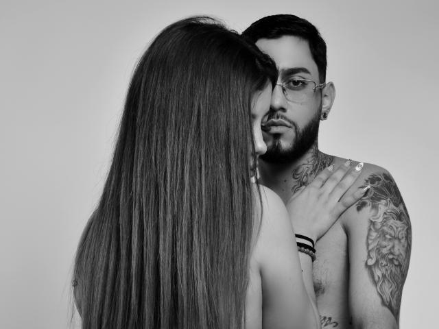 Adhara e Simon com cabelos longos, Adhara olhando para Simon, que está sem camisa, tem tatuagens, óculos e barba, ela segurando o ombro dele, a mão dela no peito dele, em preto e branco, íntimo..