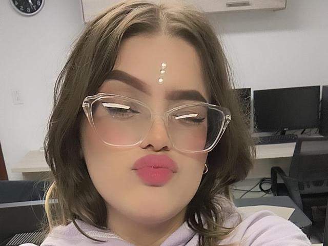Kim: Sommer mit rosigen Lippen, klaren Brillen, braunen Haaren und einem Punktmuster auf der Stirn. Ihre Nase ist leicht nach oben gebogen, und sie trägt ein helllila Oberteil..