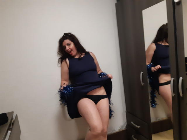 LaFrancaiseJessy, con cabello oscuro, vestida con un vestido azul marino, ropa interior negra y pompones azules, levantando el vestido para mostrar su vulva, de pie frente a un espejo, cerca de un armario de madera..