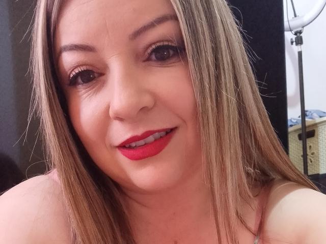 NatalyHami with red lipstick, straight blonde hair, and subtle makeup, smiling at the camera, showing her white teeth. Her pink tank top is partially visible.