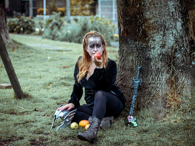 VioletaMora, met zombie-make-up, eet een appel, zittend tegen een boom, met een zwaard en helm bij zich, sinaasappels en laarzen op het gras, en ziet er "dood" uit met een "vervormd" gezicht..