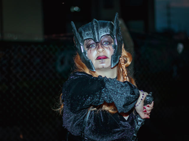 Violeta Mora, avec des cheveux roux, portant un casque Batman en métal noir, une robe noire pailletée et du rouge à lèvres, croise les bras et tient une bague, sur une photo prise de nuit..
