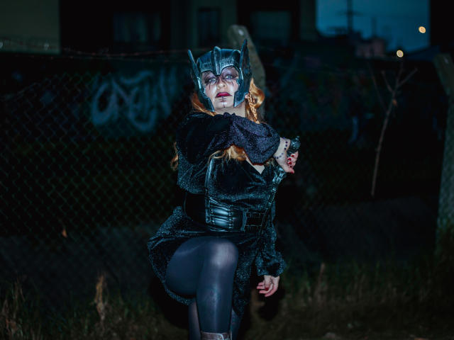 VioletaMora, dressed as a batwoman, with a black furry mask, black dress, and thigh-high stockings, squatting in a dark, grassy area, showing her ass and red lipstick.