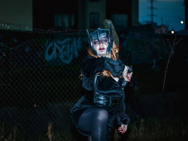 VioletaMora disfrazada de Batwoman, vistiendo un vestido negro, un corpiño y un casco, con los brazos cruzados, agachada en un entorno urbano oscuro y descuidado, y una valla de cadena detrás..