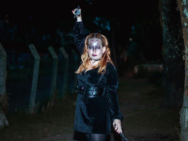 Violeta Mora, con la pelle pallida, i capelli rossi, un abito nero e una collana, che tiene un coltello nella mano destra sollevata, in piedi in una foresta oscura e inquietante di notte..