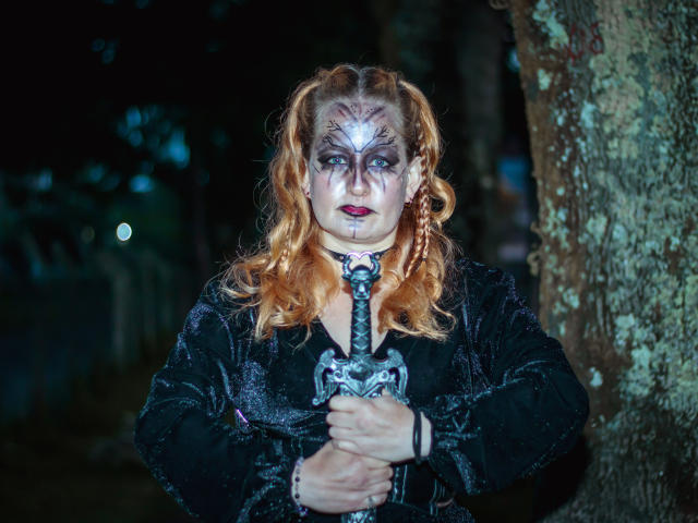 VioletaMora, avec un visage pâle et maquillé, des cheveux bouclés roux, une robe de velours noir, tenant une épée ornée, debout près d'un arbre la nuit. Sans seins apparents, maquillage sur le visage, expression sérieuse..