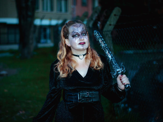 VioletaMora met bleke make-up, rode lippenstift, een zwarte jurk, een choker, die een versierde zwarte dolk vasthoudt, met golvend oranje haar, staand in een grasachtig gebied, donkere achtergrond, nacht..