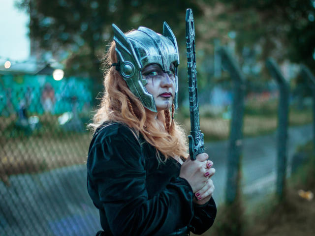 VioletaMora with silver helmet, white face paint, holding a chain sword, wearing black dress, wavy orange hair, red nails, standing by a fence in a park.