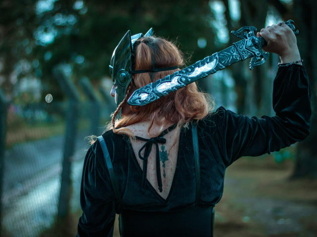 VioletaMora with wavy red hair, wearing a black velvet dress and metallic helmet, holding a large, ornate, blue-patterned sword over her shoulder, back to the camera.