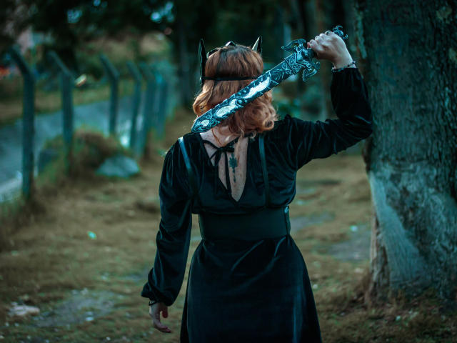 Violeta Mora, avec ses cheveux roux ondulés, sa robe noire et ses cornes de diable, tenant une épée argentée et ornée sur son épaule, debout près d'un arbre, en extérieur..