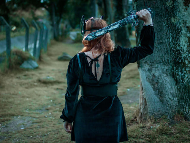 VioletaMora, con cabello rojo, vestido negro, sosteniendo una espada de plata sobre su cabeza, de pie junto a un árbol, con la espalda hacia la cámara, en un área boscosa..