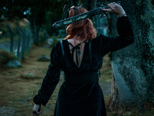 VioletaMora con i capelli rossi, vestito nero, che tiene una spada sopra la testa, in piedi di fronte a un albero, lama della spada con un motivo scuro, braccio destro sollevato, braccio sinistro in basso, albero a destra..